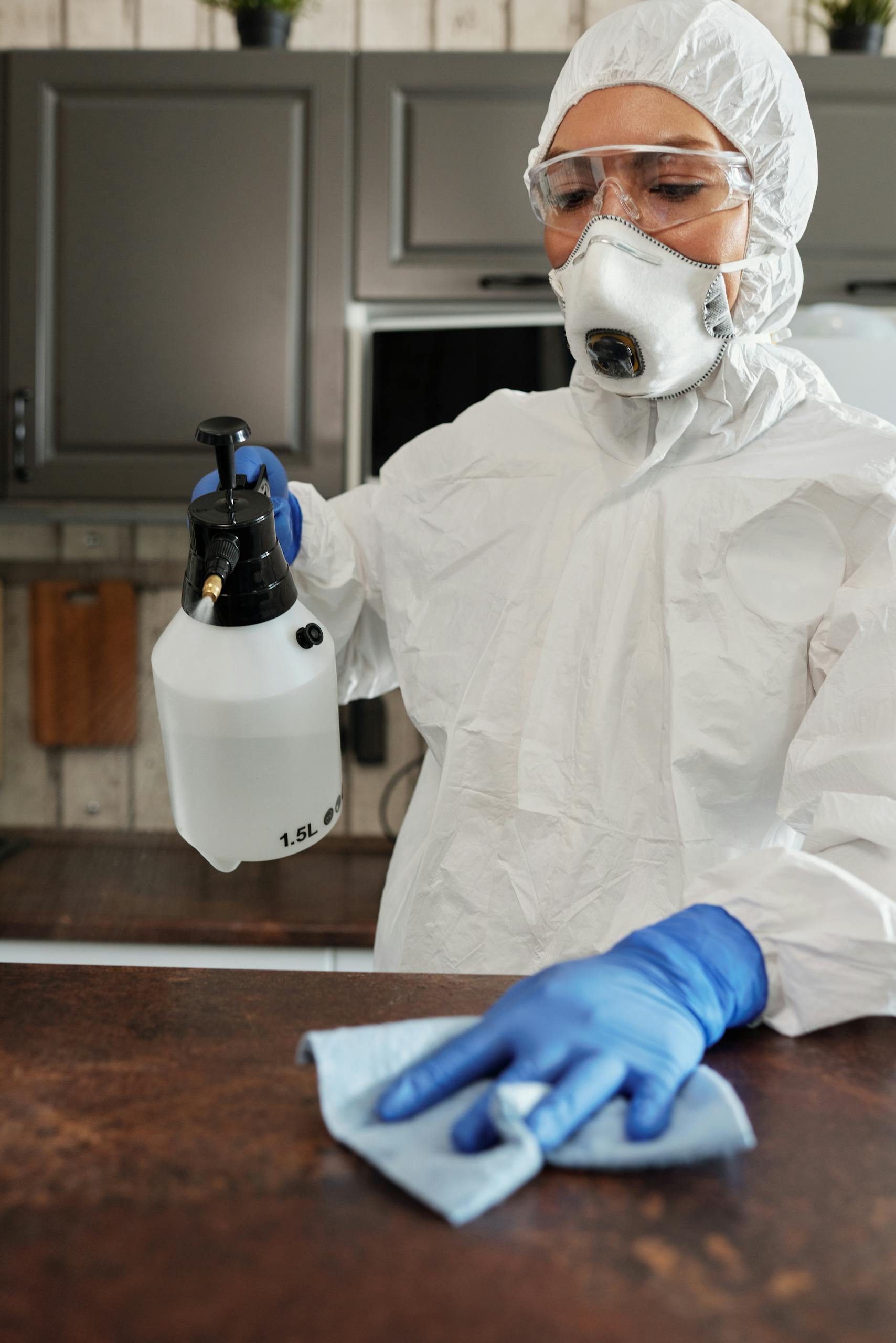 A person in protective suit cleaning a surface with disinfectant spray indoors.